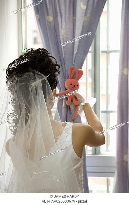 Bride facing window, tying curtain with stuffed toy, rear view