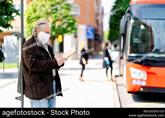 Senior man wearing protective mask and gloves reading book while waiting at bus stop, Spain