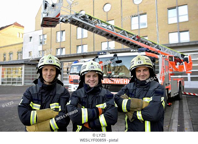 Portrait of three smiling firefighters standing on yard in front of fire engine
