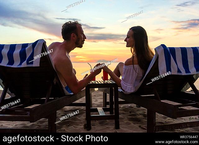 Rear view of a young couple holding hands while watching together the sunset on the dreamy beach of an exotic island, during summer vacation or honeymoon