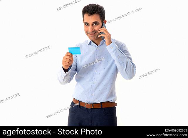 A HAPPY PROFESSIONAL LOOKING AT CAMERA WHILE HOLDING VISITING CARD AND TALKING ON MOBILE