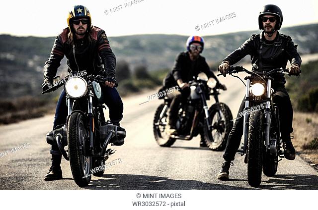 Three men wearing open face crash helmets and goggles sitting on cafe racer motorcycles on a rural road