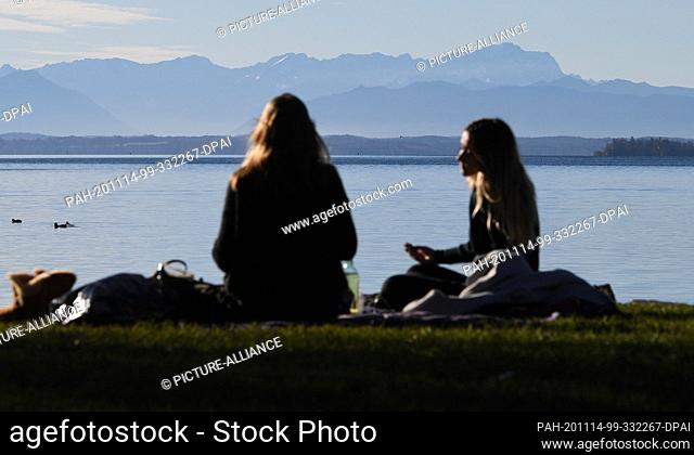 14 November 2020, Bavaria, Starnberg: Two young women are sitting in the sun at Lake Starnberg, in the background the Wetterstein mountain range with the...