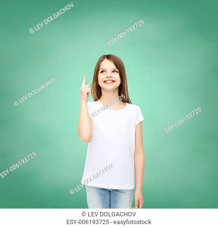 advertising, gesture, education, childhood and people - smiling little girl in white t-shirt pointing finger up over green board background