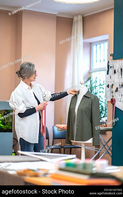 Idea. Gray-haired woman with measure in hands standing sideways to camera thoughtfully touching shirt dressed on mannequin in atelier