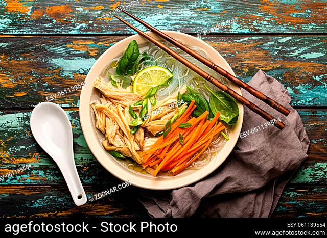 Asian soup with rice noodles, chicken and vegetables in ceramic bowl served with spoon and chopsticks on rustic wooden background from above