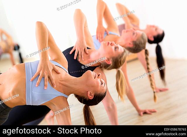 Group of young sporty attractive women in yoga studio, practicing yoga lesson with instructor, forming a line in Trikanasana asana pose