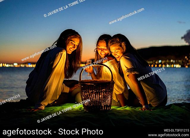 Smiling sisters looking at illuminated basket while sitting against sea during dusk