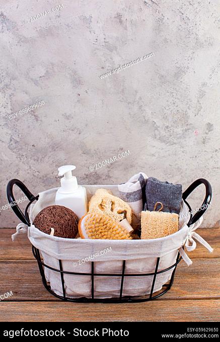 Picture of zero waste concept gift bath set in wire basket. Brush, luffah, soap, towel in black metal basket on wooden table, light grey background