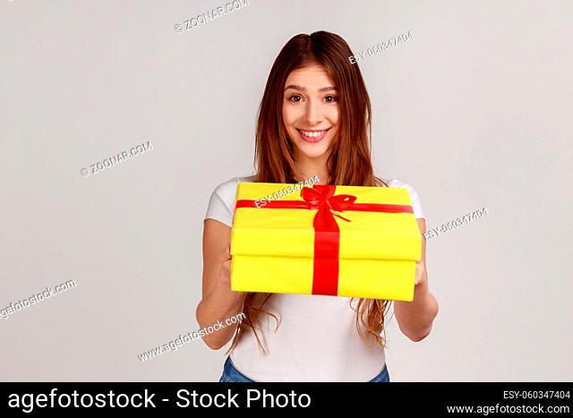 Portrait of smiling dark haired woman giving yellow wrapped present box, congratulating with holiday, wearing white casual style T-shirt