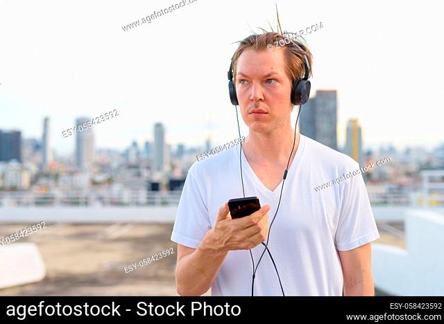 Portrait of young handsome Scandinavian man with blond hair against view of the city at rooftop of building outdoors