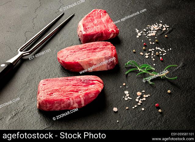 Raw beef steaks with salt, pepper, rosemary and a carving fork on a black background