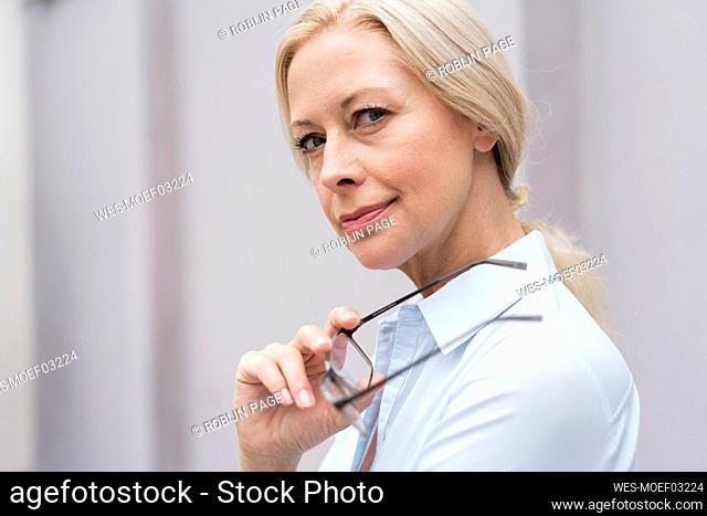 Close-up portrait of businesswoman holding eyeglasses against wall in office