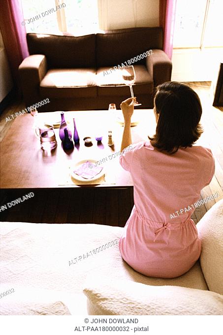 Woman sitting in living room, holding up wine glass, rear view