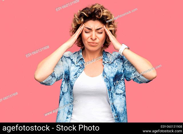 Headache, thinking or confusion. Portrait of sad young woman with curly hair in casual blue shirt standing and holding her painful head with closed eyes