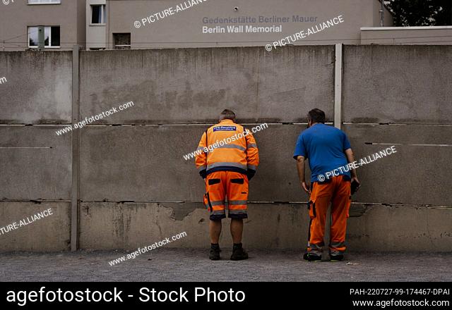 27 July 2022, Berlin: Two men look through a crack in the wall at a remnant of the former border fortifications on the grounds of the Berlin Wall Memorial on...
