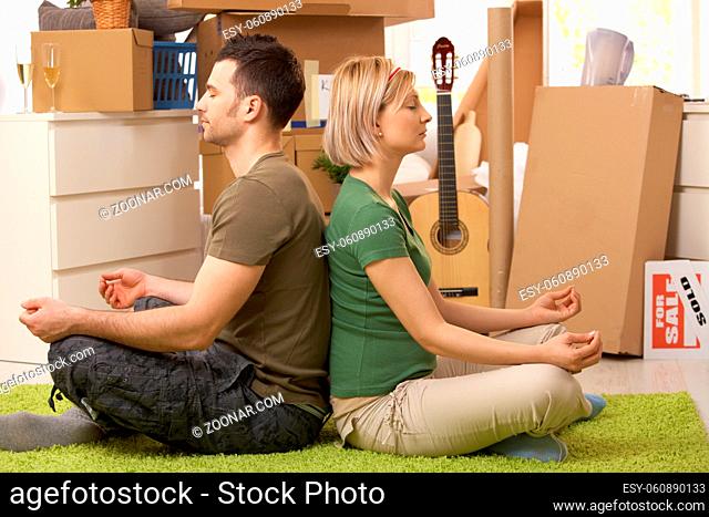 Young couple sitting in yoga meditation posture on carpet in new house, surrounded with boxes