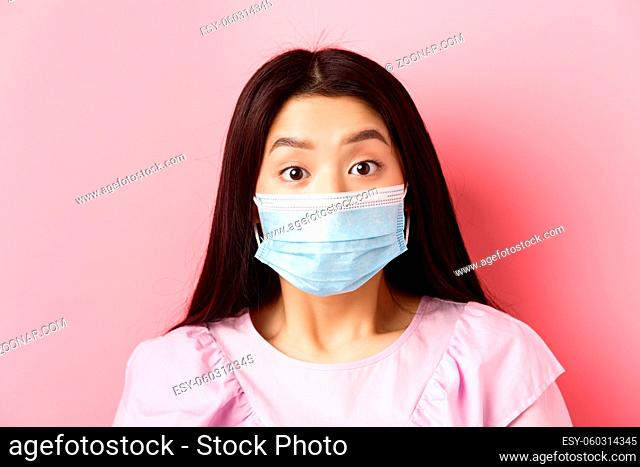Covid-19 and healthy people concept. Close-up portrait of surprised asian girl in medical mask looking excited at camera, standing against pink background