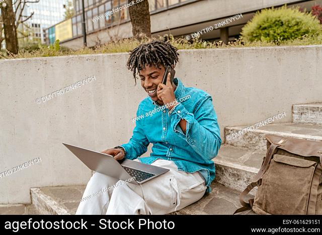 Freelancer. Young smiling dark-skinned man working on laptop talking on smartphone sitting on steps on city street on fine day
