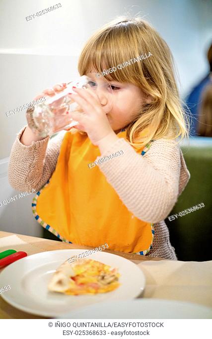portrait of three years old blonde caucasian child with orange bib drinking water in crystal glass at pizza restaurant