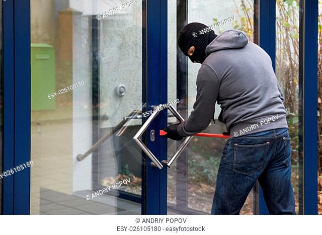 Hooded man using crowbar to open glass door