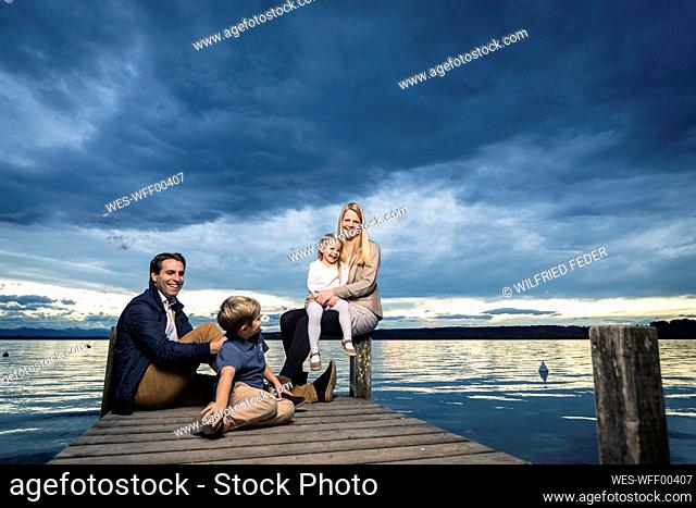 Full length of happy family sitting on jetty at Starnberger See against cloudy sky