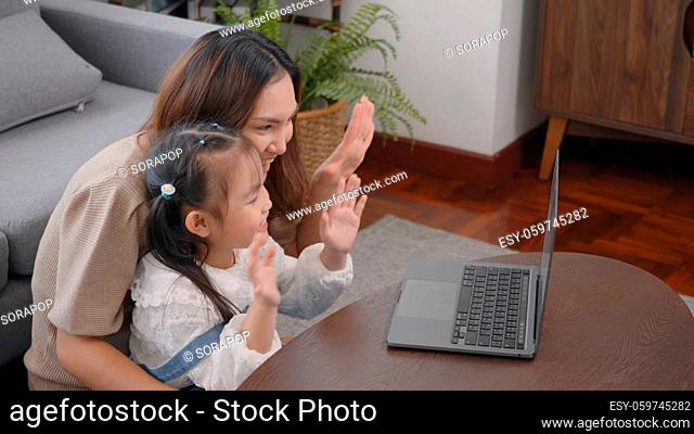 Asian young mother with laptop computer and her kid to video call camera internet online with someone in living room at home