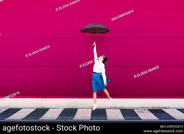 Young woman jumping with umbrella in front of a pink wall