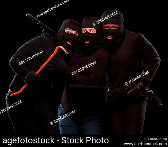 Portrait of three gangsters posing with rifle, crowbar and gun in studio. Dangerous people in balaclavas over dark grey background. Isolated on black