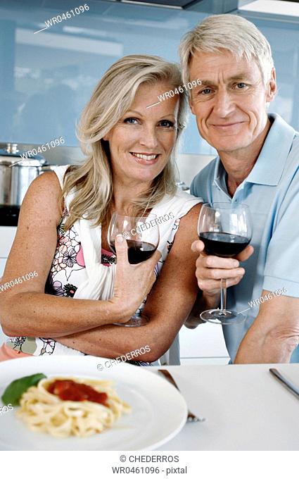 Portrait of a senior couple holding glasses of red wine and smiling at the kitchen counter