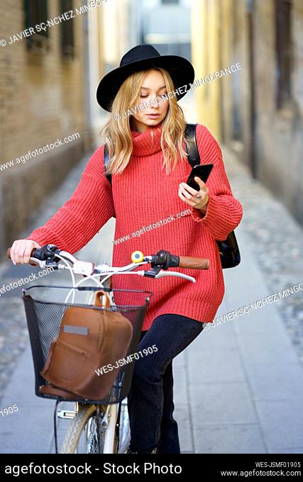 Woman wearing hat using mobile phone while standing with bicycle on footpath