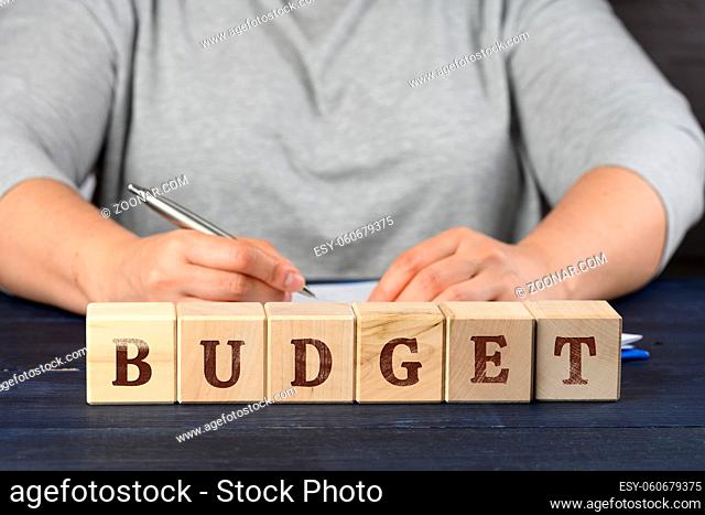 woman sits at the table and signs documents with a pen, in front of wooden cubes with the inscription budget. Audit concept, revision, cost estimate approval