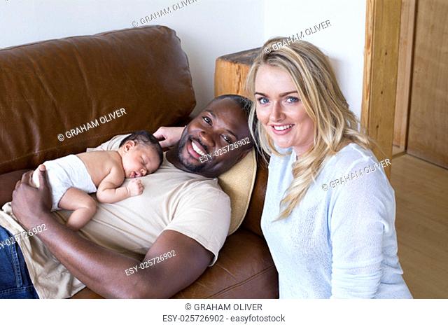 A mother and father with their newborn baby daughter at home. They are looking at the camera and smiling