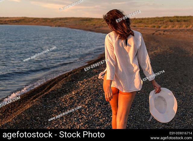 Woman walking on rocky beach with hat, rear view
