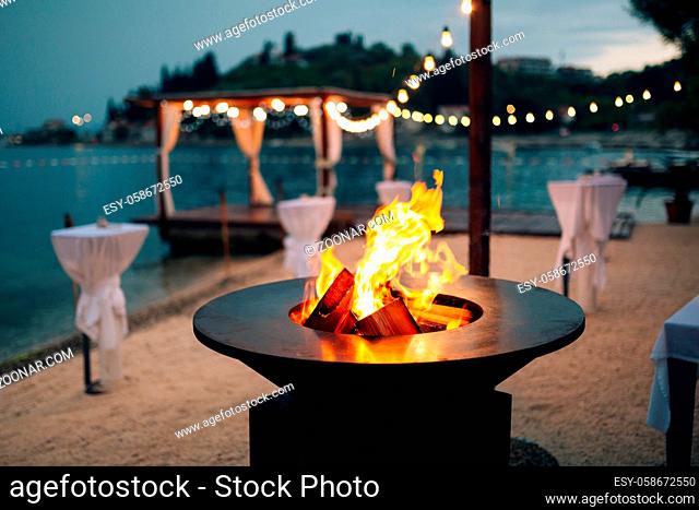 Grill with flames inside. Round table-cooking surface. On the beach, in the background of the gazebo by the water with garlands, in the twilight light