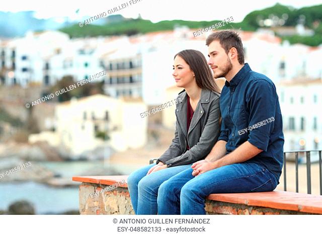 Pensive couple looking away sitting on a ledge on vacation in a coast town