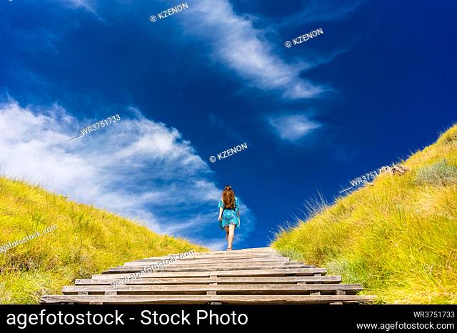Full length rear view of a young woman wearing a blue dress while climbing wooden stairs outdoors in an idyllic travel destination from Indonesia