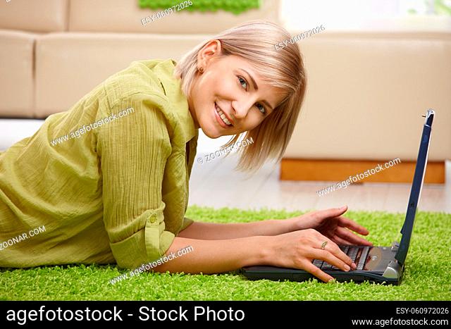 Smiling woman looking at camera, lying on floor of living room using computer