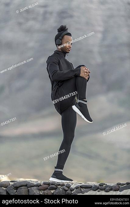 Side view of young African American man listening to music and pulling knee to chest while exercising on blurred background of countryside