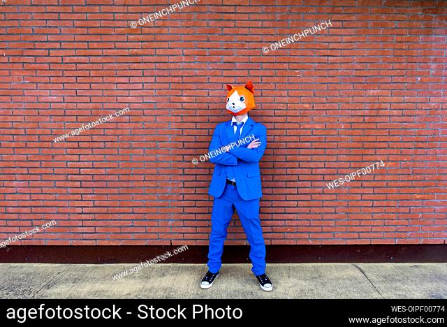Man wearing vibrant blue suit and cat mask standing with crossed arms in front of brick wall