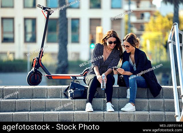 Smiling friends using mobile phone while sitting on staircase in city