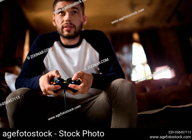 View of sportive young man having fun, playing video games. Handsome male at home, siiting on sofa, holding joystick in hands