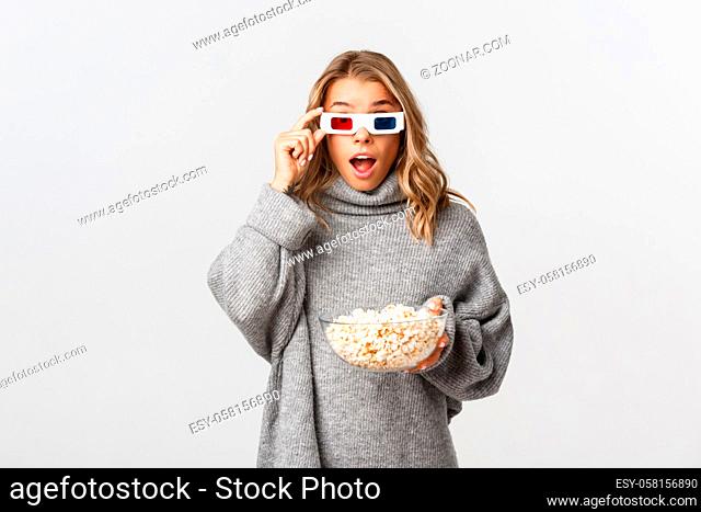Portrait of surprised cute girl in grey sweater and 3d glasses, eating popcorn, watching interesting movie, standing over white background