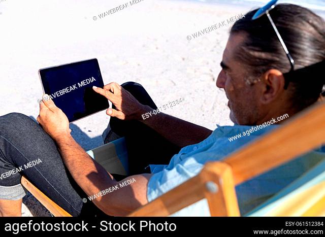 Senior Caucasian man sitting on a deck chair and using a digital tablet at the beach