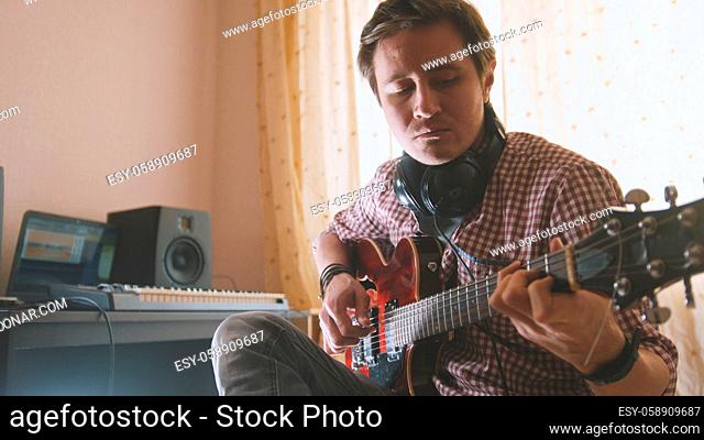 Young attractive male musician composes and records soundtrack playing the guitar, using computer, focus on face and hands, close-up