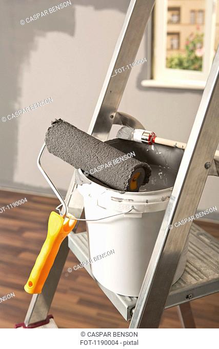 A ladder with paint roller and paintbrush leaning on paint bucket