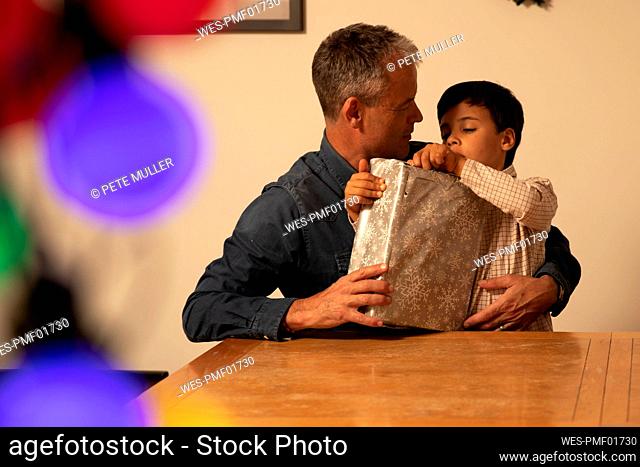 Father with curious son unwrapping Christmas present on table at home