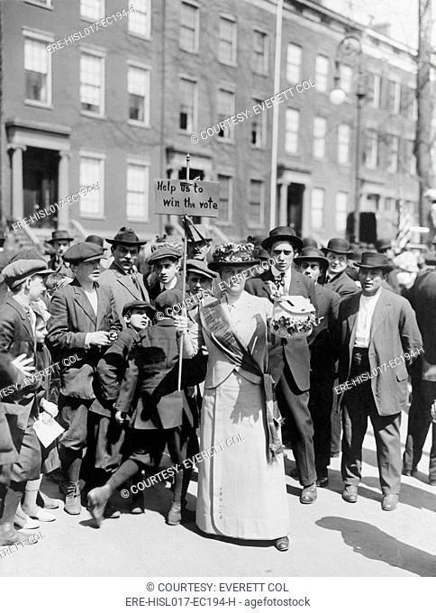 Women identified as Mrs. Suffern, is surrounded by a crowd of men and boys, while she holds a home-made banner in women suffragist parade 'Help us to win the...