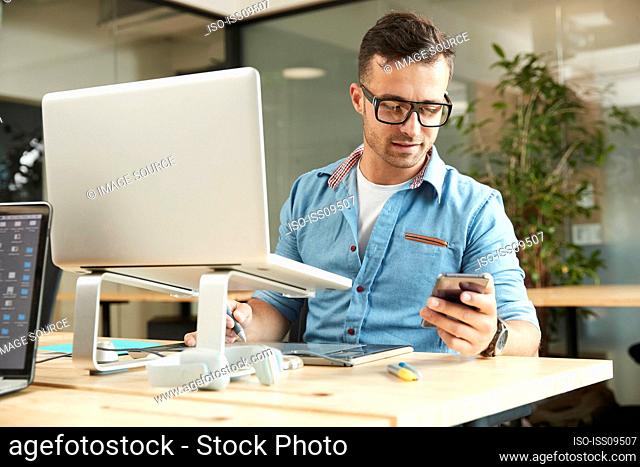 Young man holding smart phone by laptop