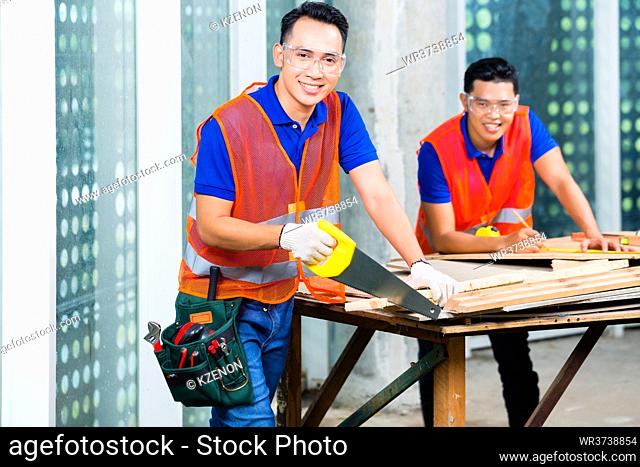 Asian Indonesian builder or craftsman sawing with a saw a wood board of a tower building or construction site wearing protection glasses and gloves
